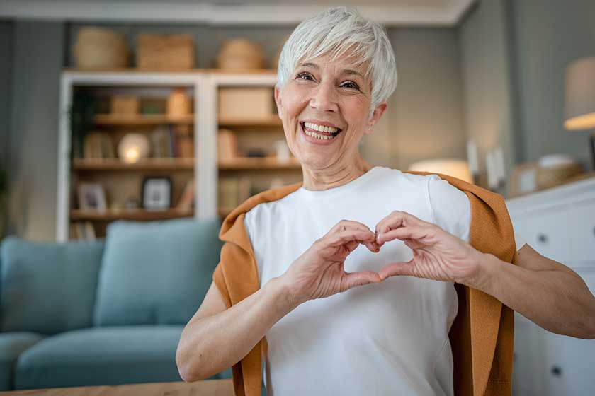 Healthy senior caucasian woman showing hart hand gesture health love happy smile while sit at home looking to the camera smiling copy space